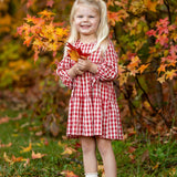 A young girl smiles outdoors, holding orange autumn leaves and wearing the Mabel and Honey Tis' the Season Dress, surrounded by colorful fall foliage.