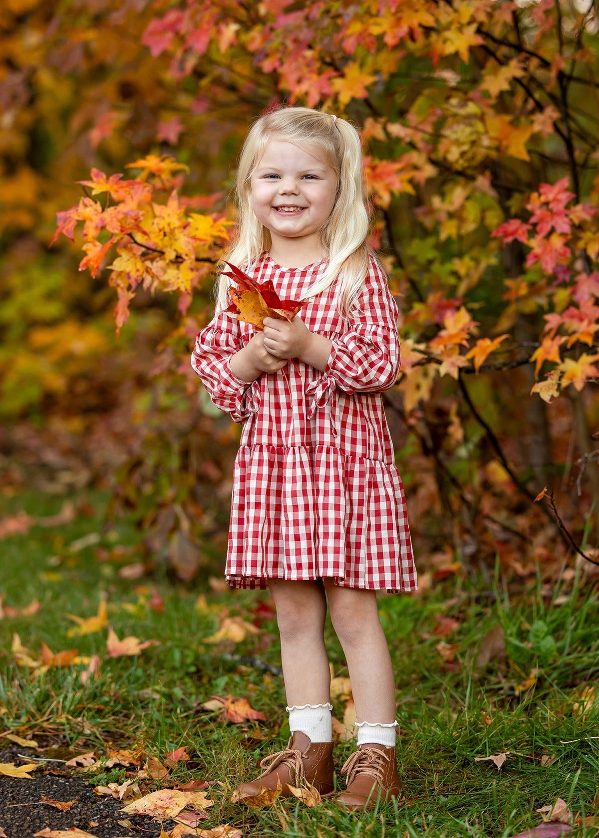 A young girl smiles outdoors, holding orange autumn leaves and wearing the Mabel and Honey Tis' the Season Dress, surrounded by colorful fall foliage.
