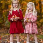 Two girls smile outdoors among fall foliage. One wears the Mabel and Honey Tis' the Season Dress with white boots, while the other sports a red gingham dress and brown shoes. Both hold autumn leaves in their hands.