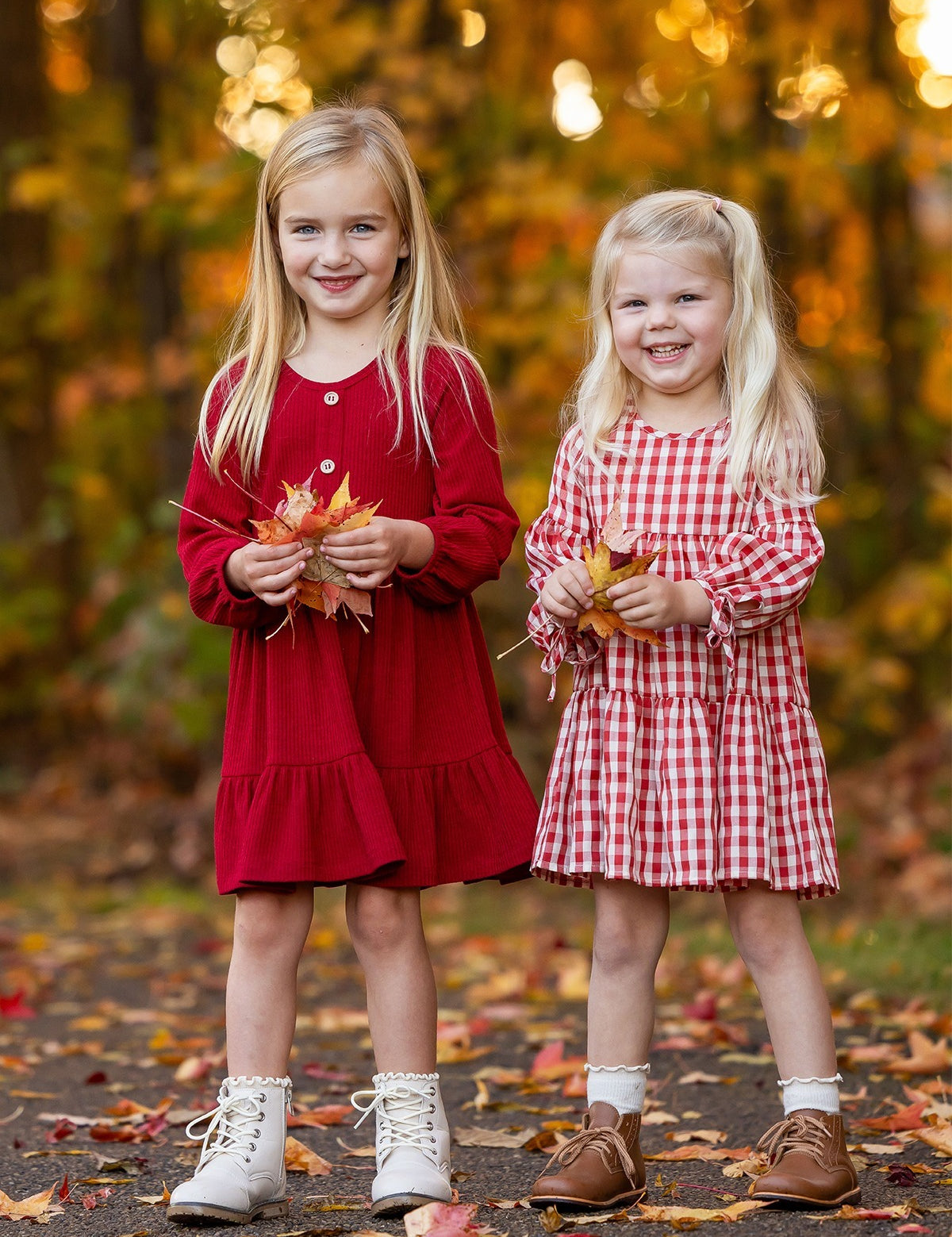 Two girls smile outdoors among fall foliage. One wears the Mabel and Honey Tis' the Season Dress with white boots, while the other sports a red gingham dress and brown shoes. Both hold autumn leaves in their hands.