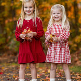 Two girls smile outdoors among fall foliage. One wears the Mabel and Honey Tis' the Season Dress with white boots, while the other sports a red gingham dress and brown shoes. Both hold autumn leaves in their hands.