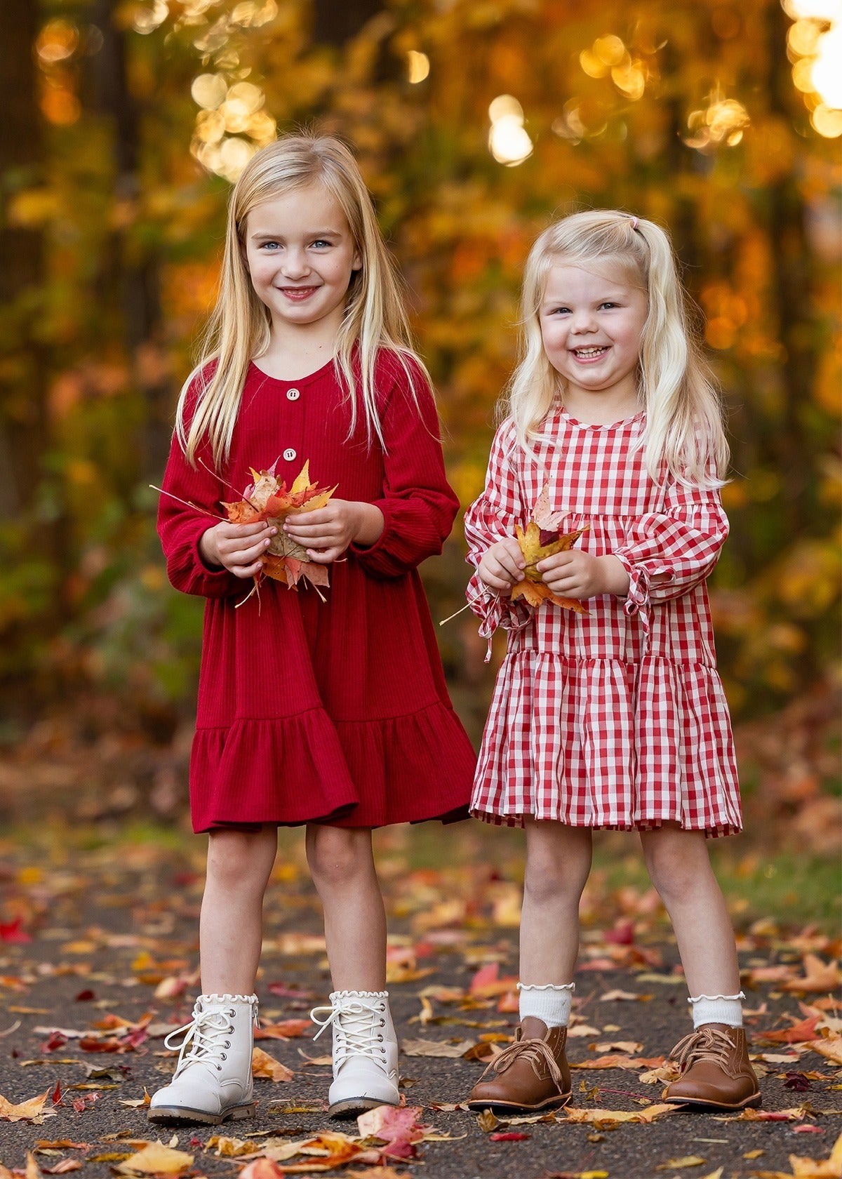 Two girls smile outdoors among fall foliage. One wears the Mabel and Honey Tis' the Season Dress with white boots, while the other sports a red gingham dress and brown shoes. Both hold autumn leaves in their hands.