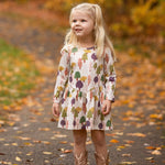 A young girl with long blonde hair smiles on a leafy autumn path, wearing the Treehouse Dress by Mabel and Honey. The nature-inspired style adds playful elegance as she blends into the golden fall scenery.