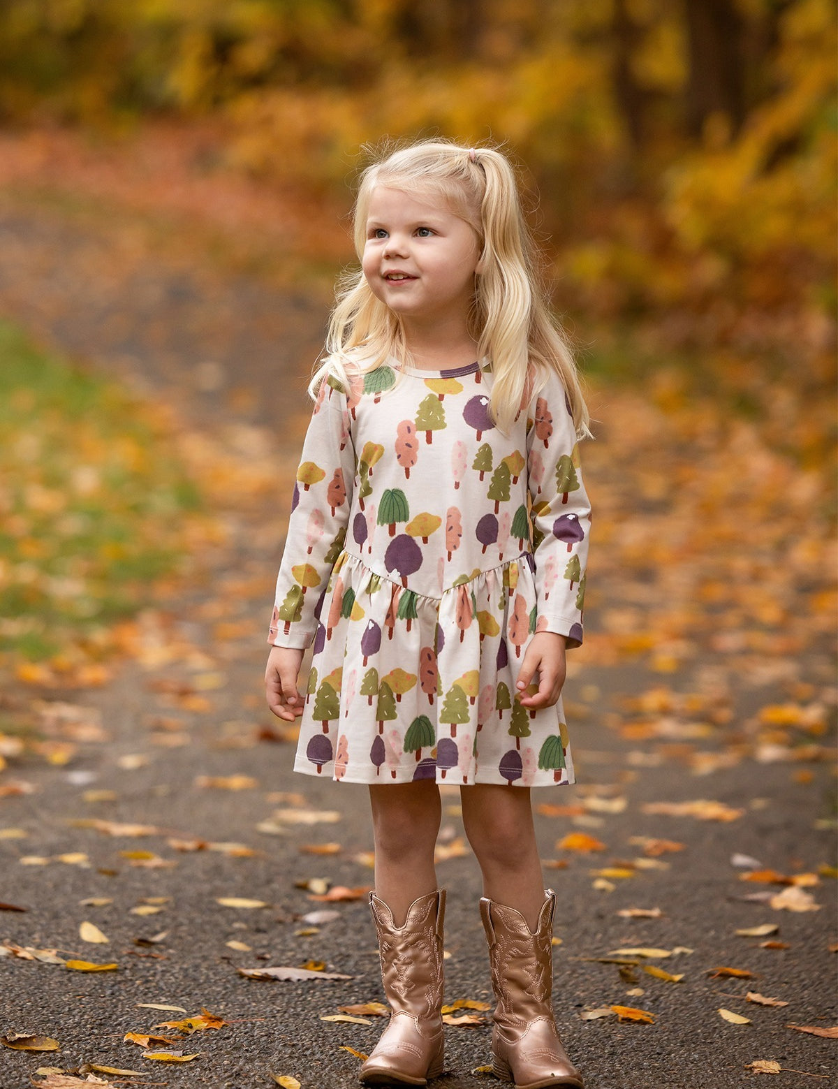 A young girl with long blonde hair smiles on a leafy autumn path, wearing the Treehouse Dress by Mabel and Honey. The nature-inspired style adds playful elegance as she blends into the golden fall scenery.