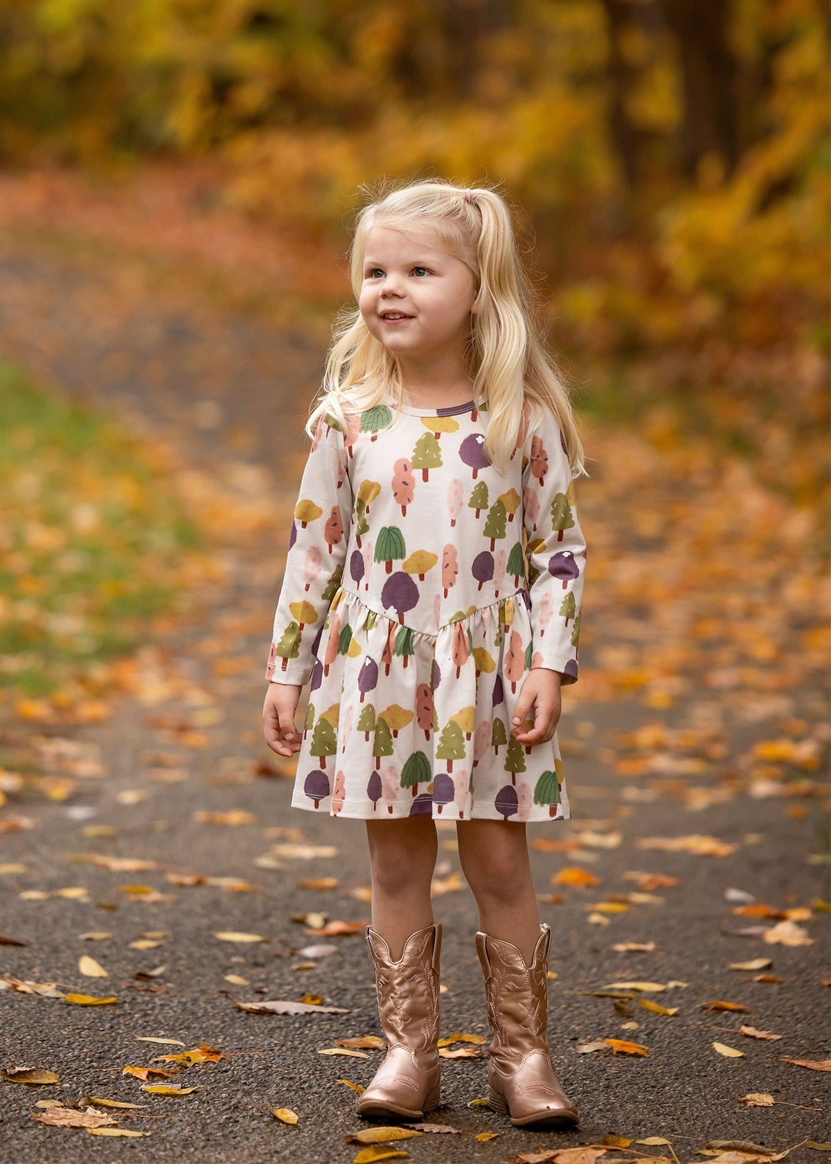 A young girl with long blonde hair smiles on a leafy autumn path, wearing the Treehouse Dress by Mabel and Honey. The nature-inspired style adds playful elegance as she blends into the golden fall scenery.