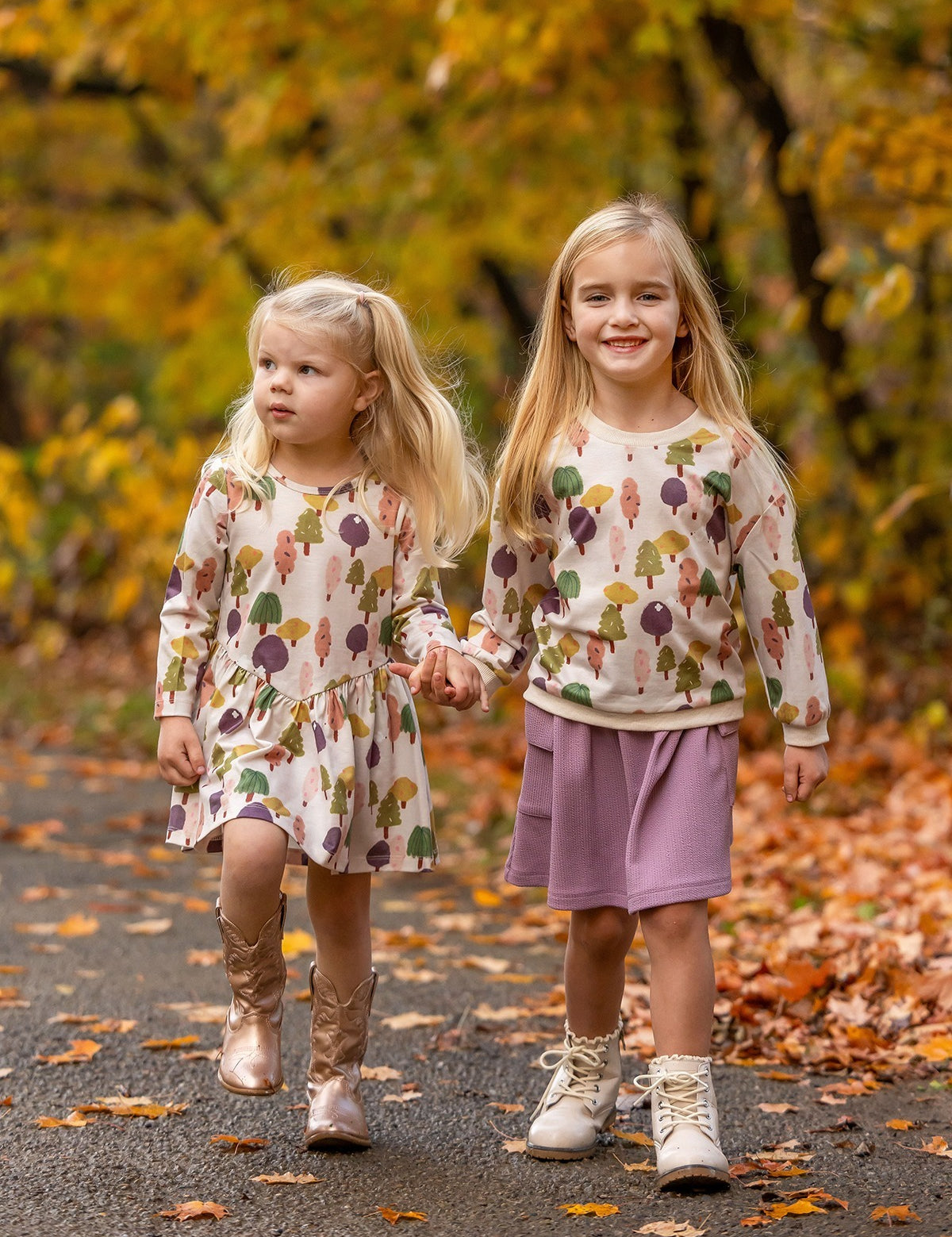 Two young girls stroll hand in hand on a leafy autumn path, dressed in matching Mabel and Honey Treehouse Dresses. One smiles at the camera as they’re surrounded by golden fall foliage and nature-inspired style.