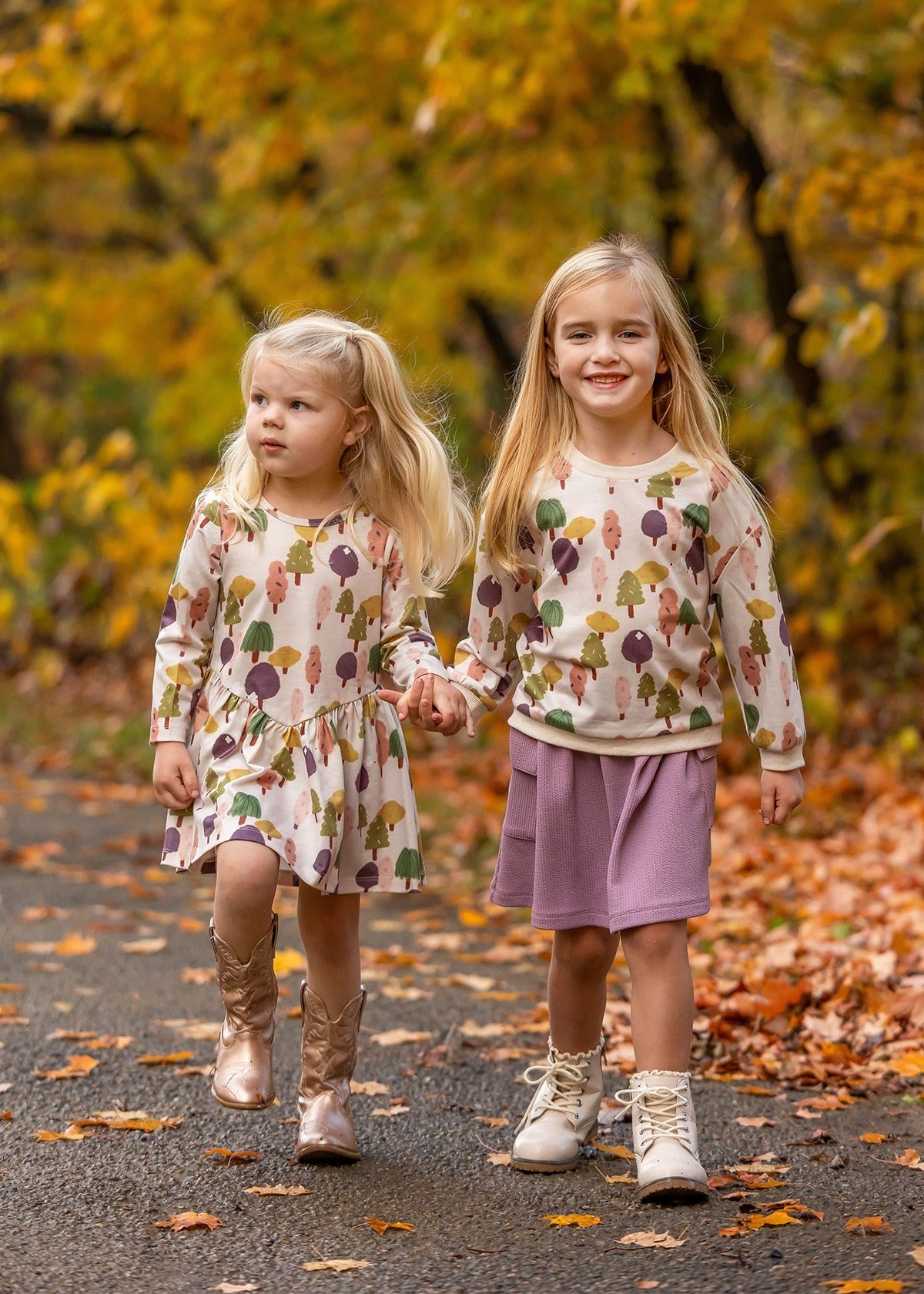 Two young girls stroll hand in hand on a leafy autumn path, dressed in matching Mabel and Honey Treehouse Dresses. One smiles at the camera as they’re surrounded by golden fall foliage and nature-inspired style.