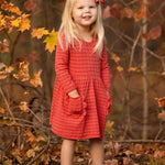 A young girl with long blonde hair, wearing the Mabel and Honey Tea Party Red Gingham Dress and tan boots, stands smiling outdoors among autumn leaves with her hands in her pockets—capturing everyday elegance.