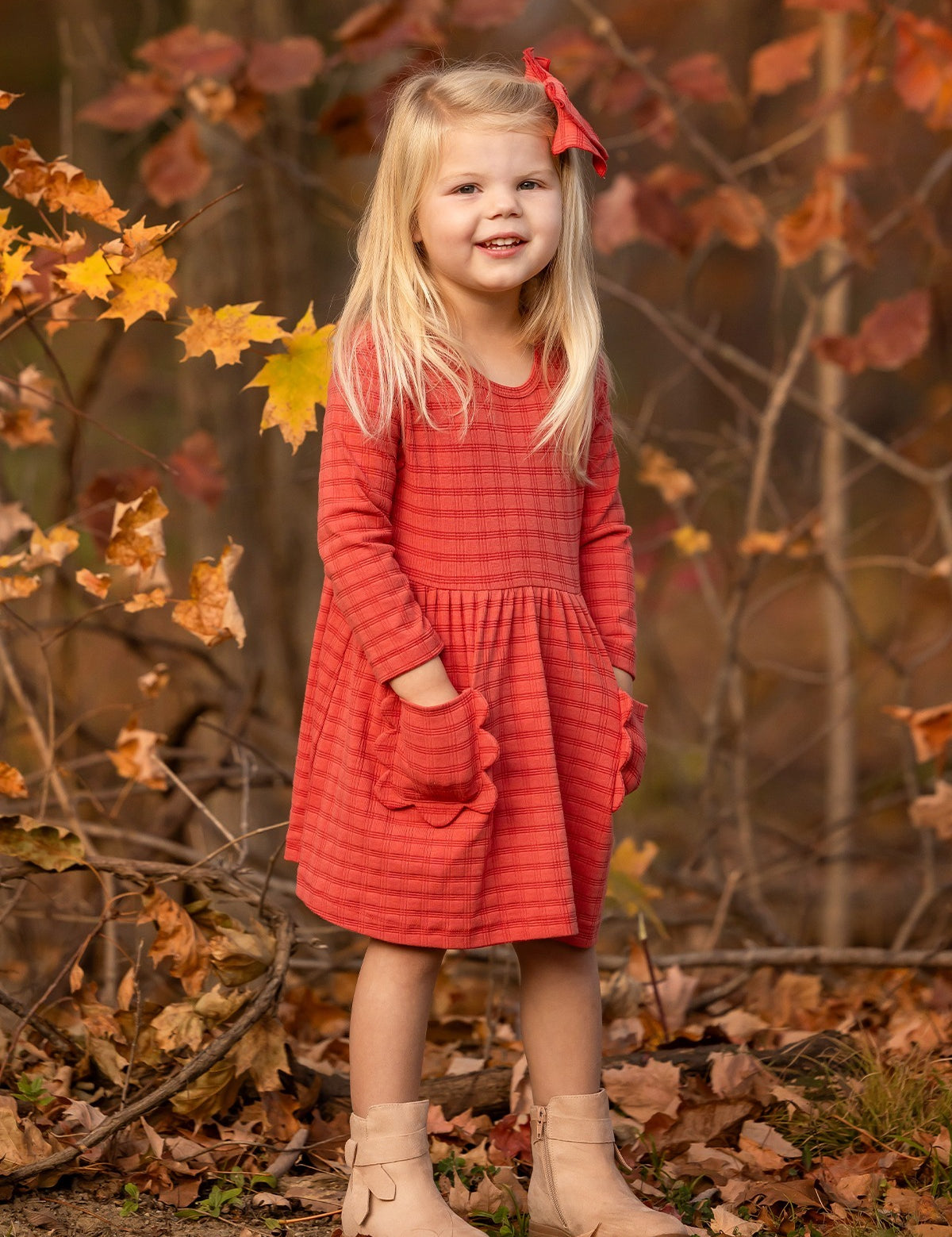 A young girl with long blonde hair, wearing the Mabel and Honey Tea Party Red Gingham Dress and tan boots, stands smiling outdoors among autumn leaves with her hands in her pockets—capturing everyday elegance.