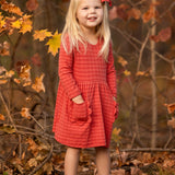 A young girl with long blonde hair, wearing the Mabel and Honey Tea Party Red Gingham Dress and tan boots, stands smiling outdoors among autumn leaves with her hands in her pockets—capturing everyday elegance.