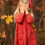 A young girl with long blonde hair, wearing the Mabel and Honey Tea Party Red Gingham Dress and a matching bow, stands outdoors by autumn leaves, shyly covering her face—a sweet display of everyday elegance.