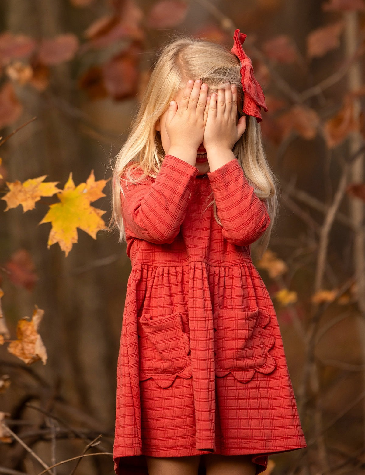 A young girl with long blonde hair, wearing the Mabel and Honey Tea Party Red Gingham Dress and a matching bow, stands outdoors by autumn leaves, shyly covering her face—a sweet display of everyday elegance.