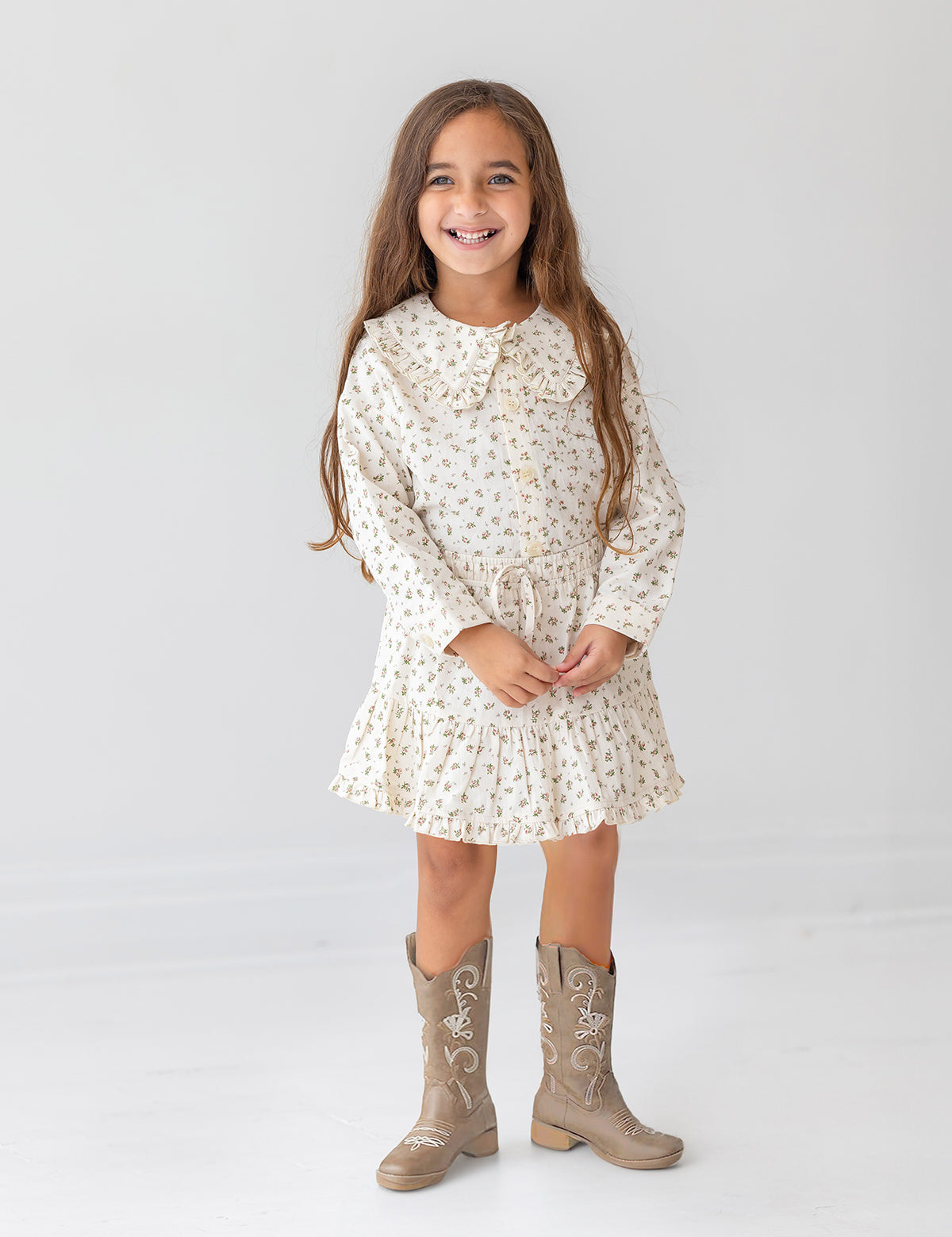 A young girl with long brown hair smiles against a white background, wearing the Mabel and Honey Juliette Two Piece Set and tan cowboy boots.