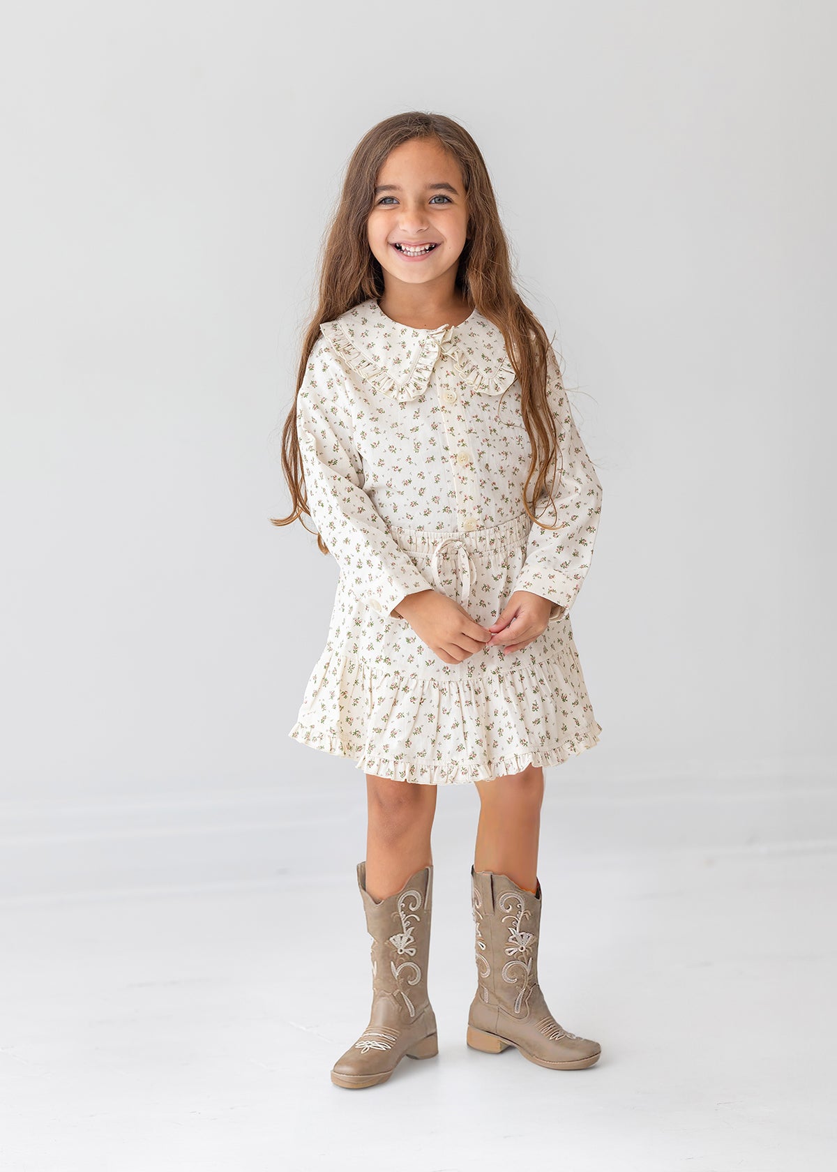 A young girl with long brown hair smiles against a white background, wearing the Mabel and Honey Juliette Two Piece Set and tan cowboy boots.