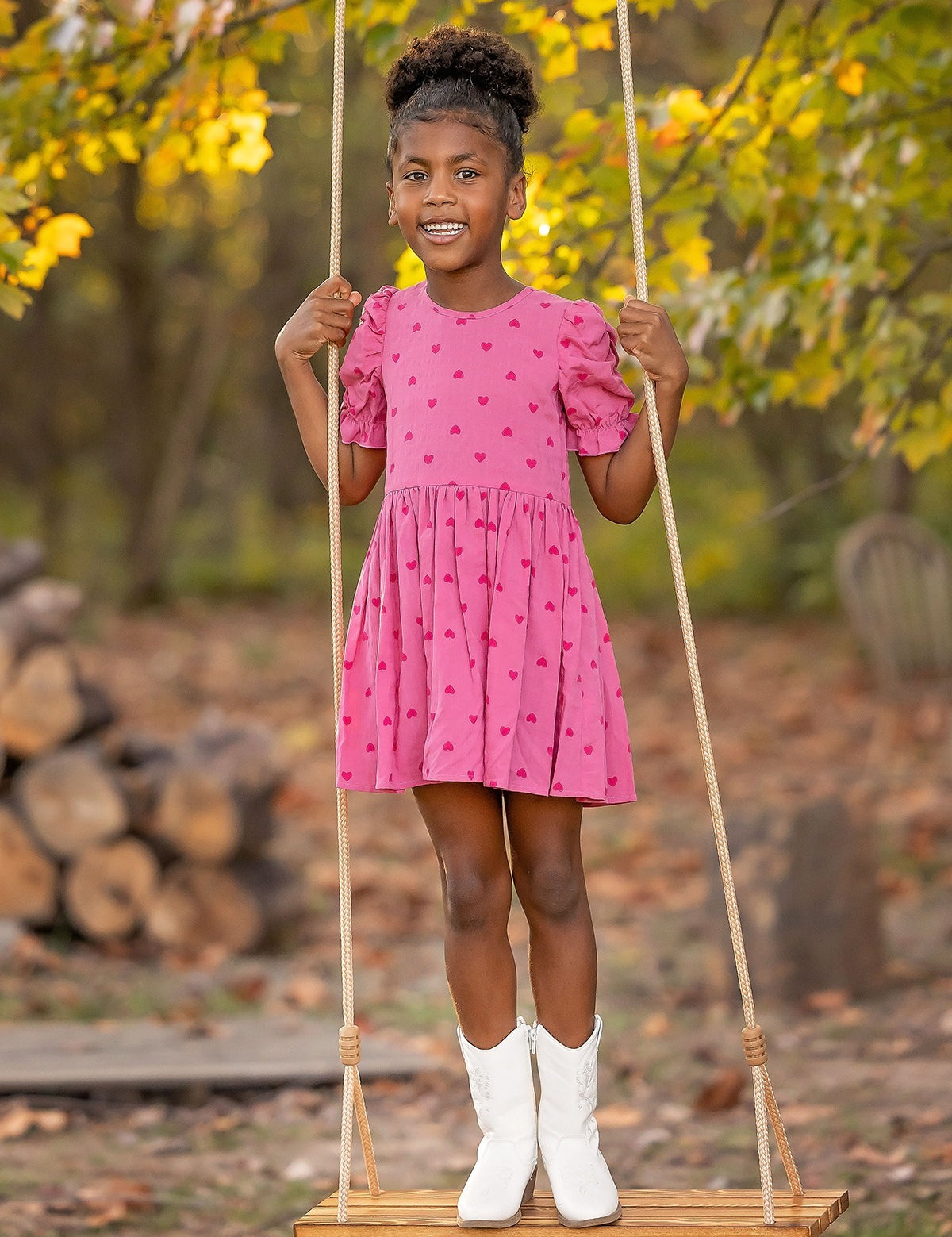 A young girl beams on a wooden swing outdoors, wearing the Mabel and Honey Perfect in Pink Hearts Short Sleeve Dress, paired with white boots amid autumn trees and stacked firewood.