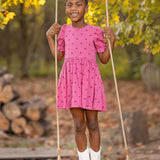 A young girl beams on a wooden swing outdoors, wearing the Mabel and Honey Perfect in Pink Hearts Short Sleeve Dress, paired with white boots amid autumn trees and stacked firewood.