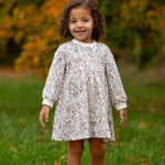 A young child with curly hair stands on green grass, smiling in the Mabel and Honey Meadow Dress—ideal for comfortable everyday wear. Blurred greenery and orange foliage in the background suggest an autumn setting.