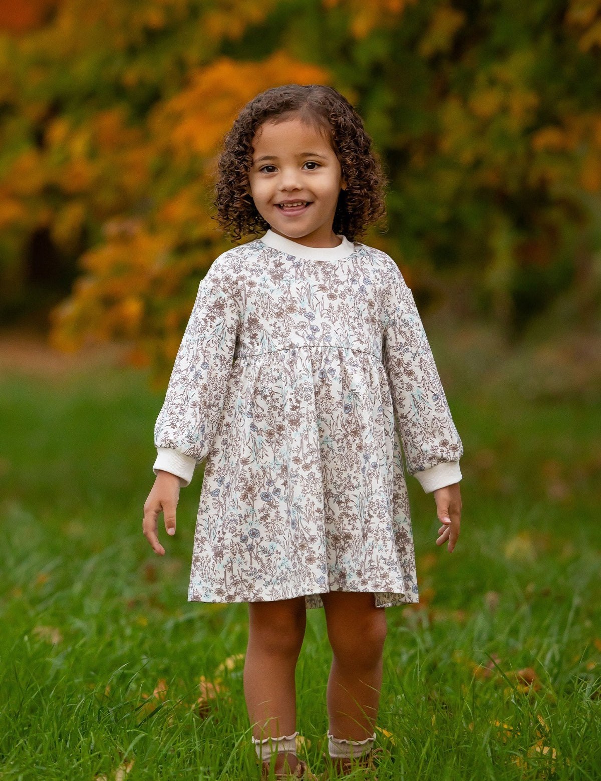 A young child with curly hair stands on green grass, smiling in the Mabel and Honey Meadow Dress—ideal for comfortable everyday wear. Blurred greenery and orange foliage in the background suggest an autumn setting.