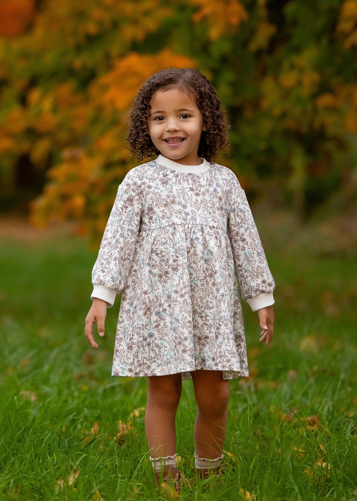 A young child with curly hair stands on green grass, smiling in the Mabel and Honey Meadow Dress—ideal for comfortable everyday wear. Blurred greenery and orange foliage in the background suggest an autumn setting.