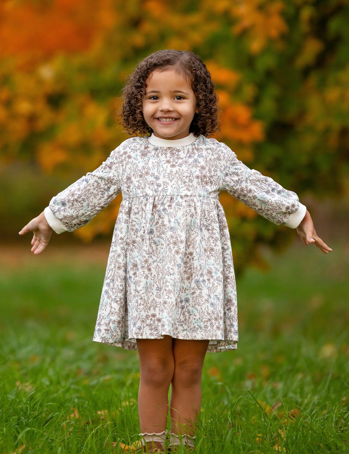 A young girl with curly hair stands on grass, smiling with her arms outstretched. She wears the Meadow Dress by Mabel and Honey, a comfortable floral girls dress. The blurred green and orange foliage hints at autumn.