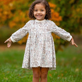 A young girl with curly hair stands on grass, smiling with her arms outstretched. She wears the Meadow Dress by Mabel and Honey, a comfortable floral girls dress. The blurred green and orange foliage hints at autumn.