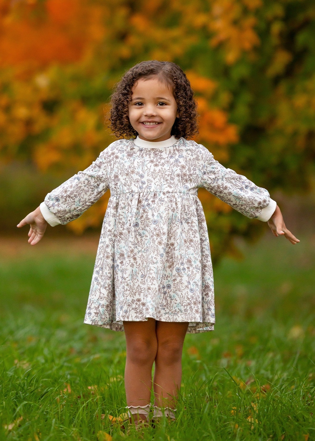 A young girl with curly hair stands on grass, smiling with her arms outstretched. She wears the Meadow Dress by Mabel and Honey, a comfortable floral girls dress. The blurred green and orange foliage hints at autumn.