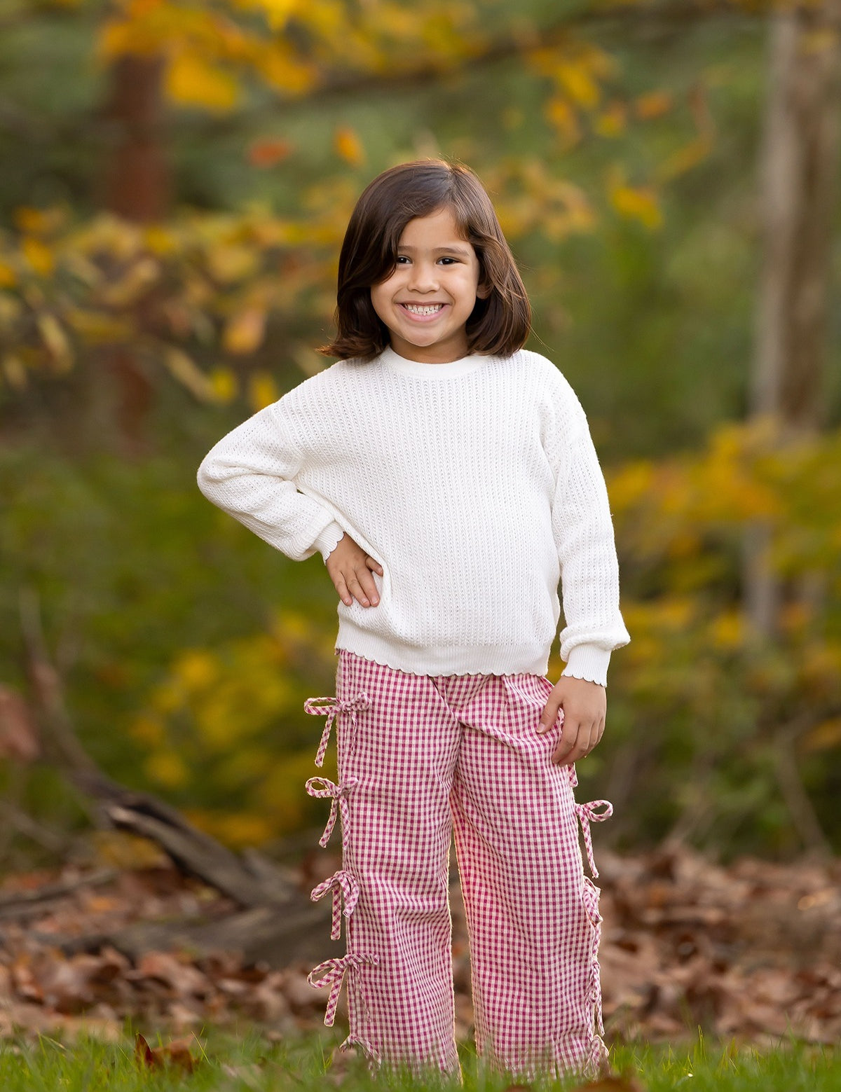 A young child with shoulder-length brown hair smiles outdoors in a white sweater and Mabel and Honey's Bow-tastic Pant, featuring an elastic waistband, surrounded by autumn leaves and greenery.