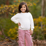 A young child with shoulder-length brown hair smiles outdoors in a white sweater and Mabel and Honey's Bow-tastic Pant, featuring an elastic waistband, surrounded by autumn leaves and greenery.