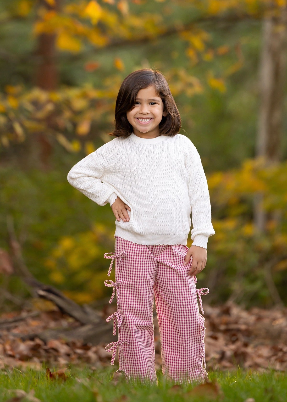 A young child with shoulder-length brown hair smiles outdoors in a white sweater and Mabel and Honey's Bow-tastic Pant, featuring an elastic waistband, surrounded by autumn leaves and greenery.