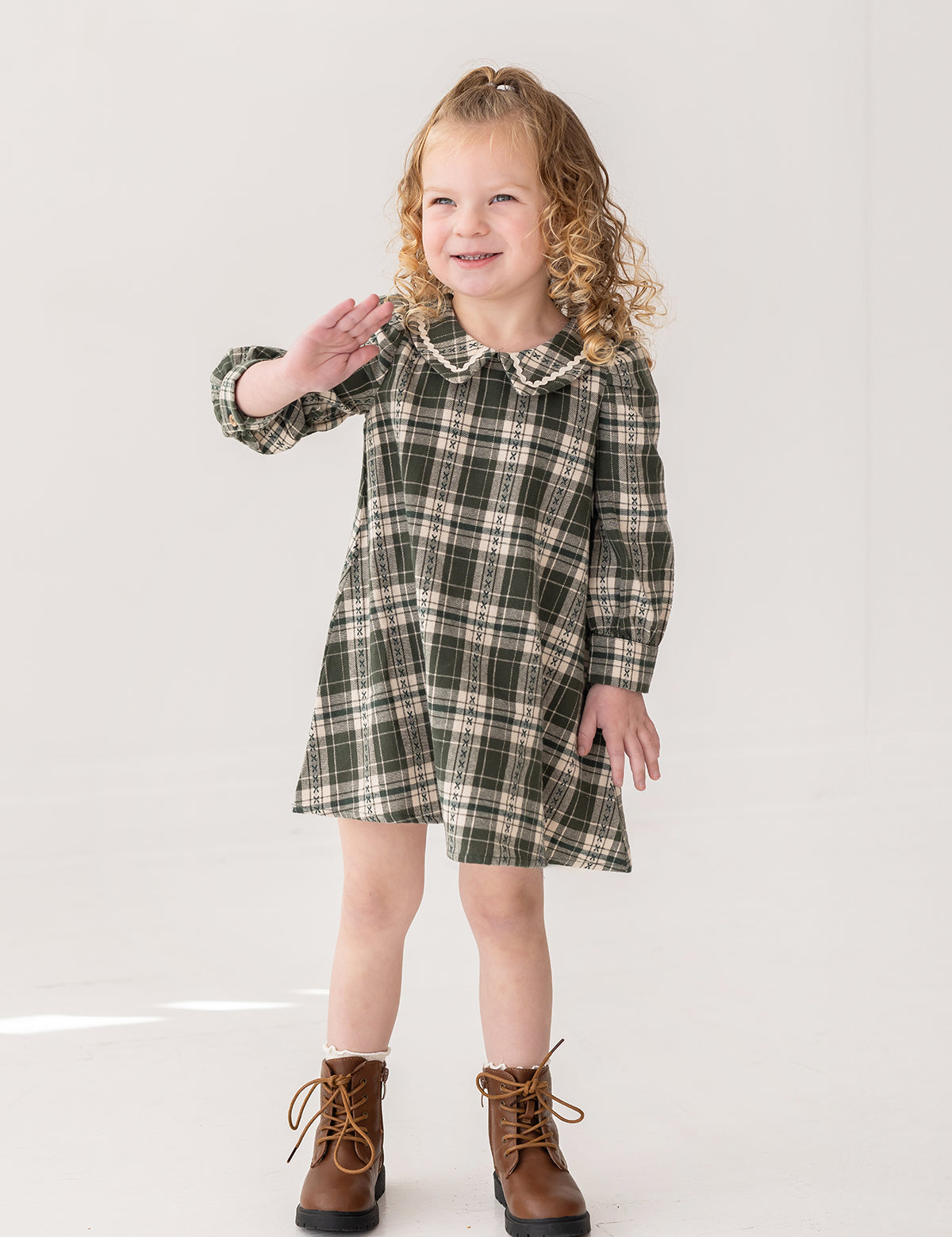 A young girl with curly blonde hair smiles and waves while modeling the Mabel and Honey Plaid & Pine Dress against a plain white background.