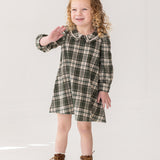 A young girl with curly blonde hair smiles and waves while modeling the Mabel and Honey Plaid & Pine Dress against a plain white background.