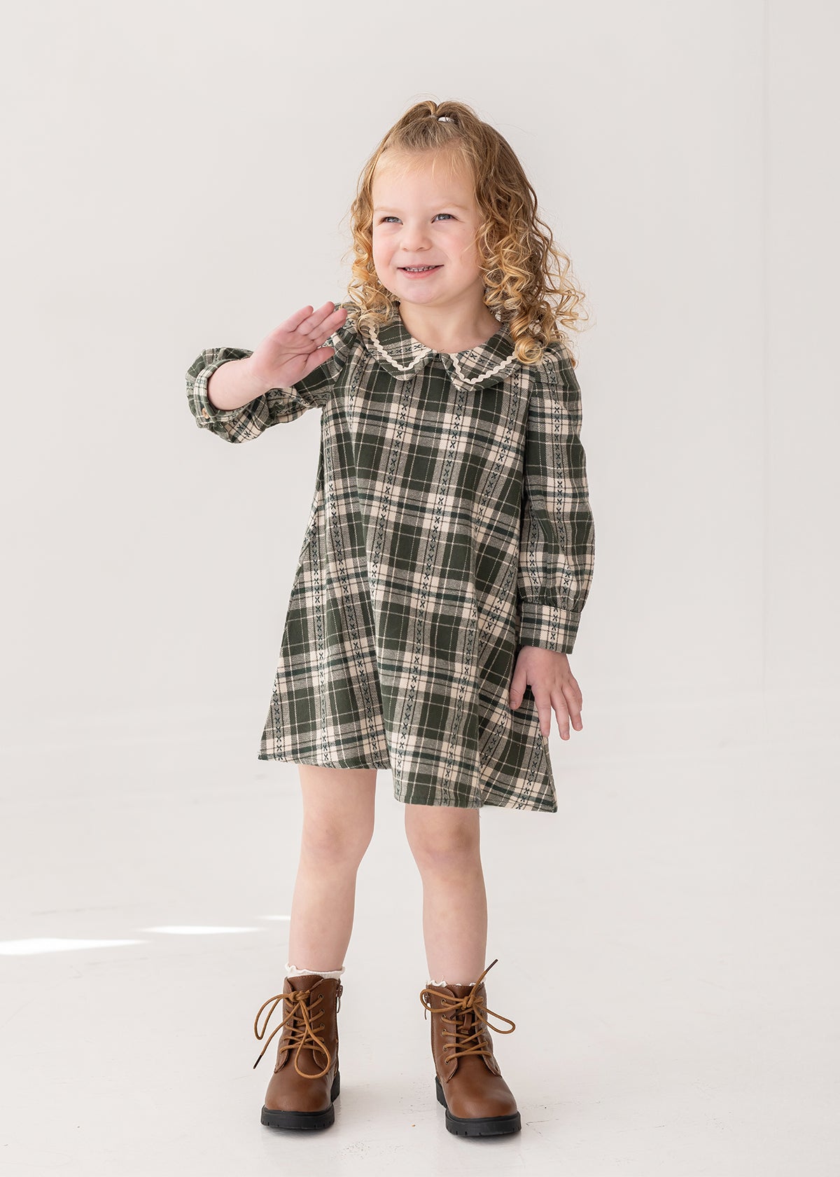 A young girl with curly blonde hair smiles and waves while modeling the Mabel and Honey Plaid & Pine Dress against a plain white background.