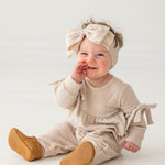 A smiling baby girl in a Mabel and Honey Cinnamon Sugar Romper with bow details and a matching headband sits on the floor, touching her mouth while wearing tan shoes against a plain white background.