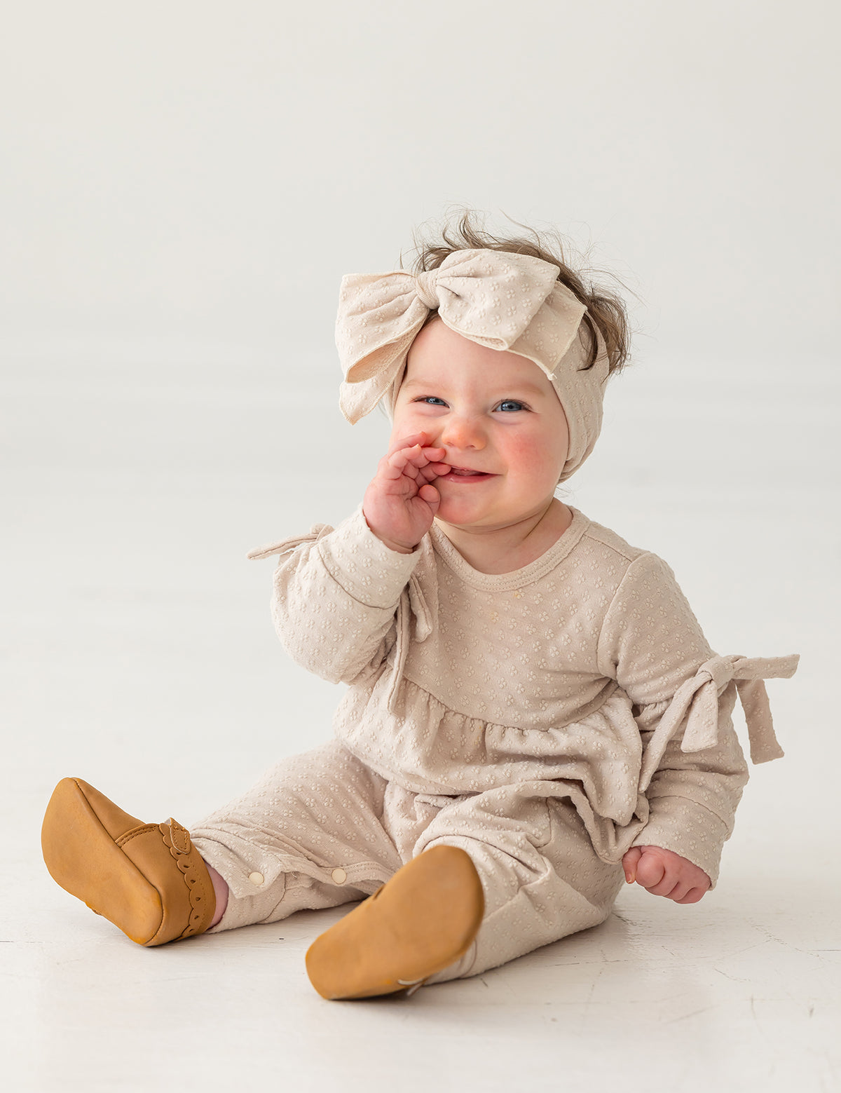 A smiling baby girl in a Mabel and Honey Cinnamon Sugar Romper with bow details and a matching headband sits on the floor, touching her mouth while wearing tan shoes against a plain white background.