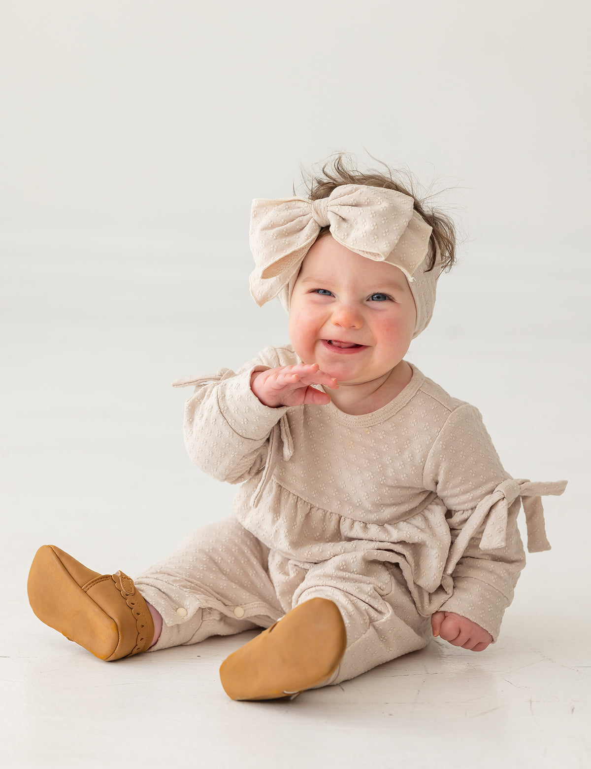 A smiling baby girl sits on a white floor against a plain background, wearing the Mabel and Honey Cinnamon Sugar Romper with bow-detailed sleeves, a matching large bow headband, and brown shoes.