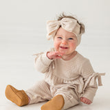 A smiling baby girl sits on a white floor against a plain background, wearing the Mabel and Honey Cinnamon Sugar Romper with bow-detailed sleeves, a matching large bow headband, and brown shoes.