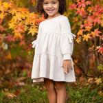 A young girl with curly hair smiles outdoors on green grass, wearing the Mabel and Honey Cinnamon Sugar Dress with a tiered silhouette and brown boots, standing in front of autumn trees with vibrant orange and yellow leaves.