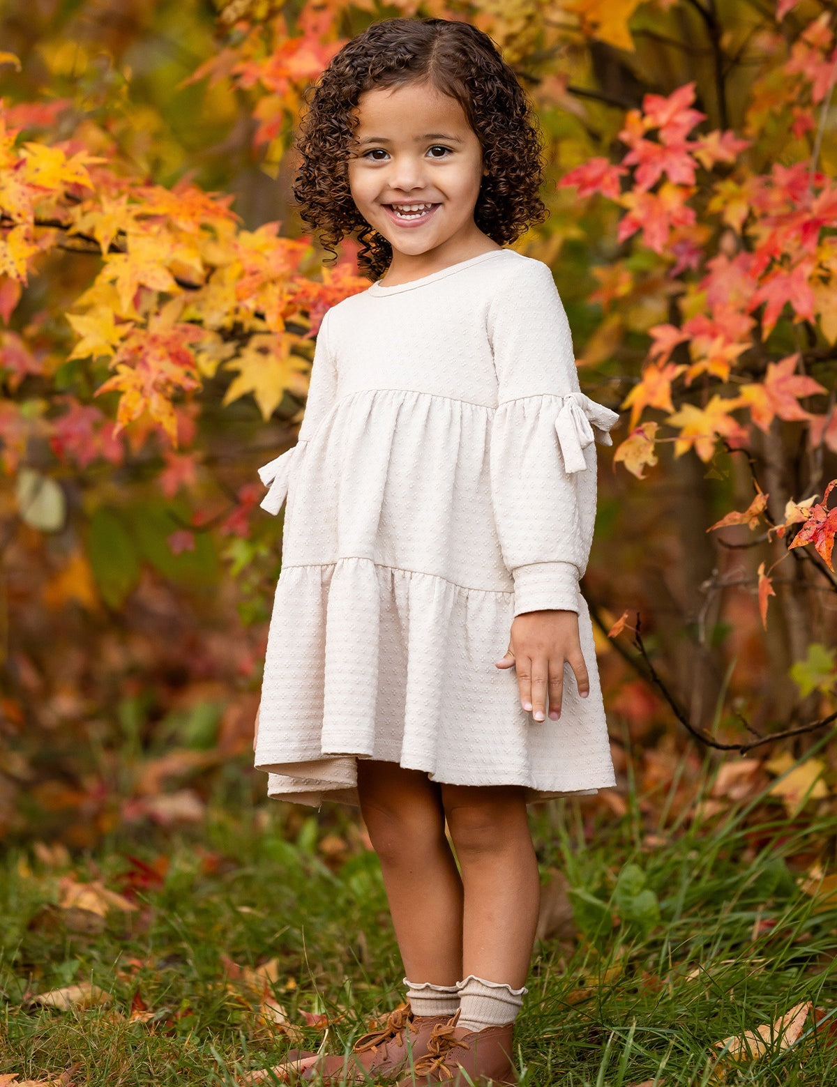 A young girl with curly hair smiles outdoors on green grass, wearing the Mabel and Honey Cinnamon Sugar Dress with a tiered silhouette and brown boots, standing in front of autumn trees with vibrant orange and yellow leaves.
