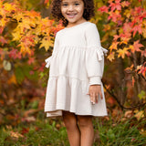 A young girl with curly hair smiles outdoors on green grass, wearing the Mabel and Honey Cinnamon Sugar Dress with a tiered silhouette and brown boots, standing in front of autumn trees with vibrant orange and yellow leaves.