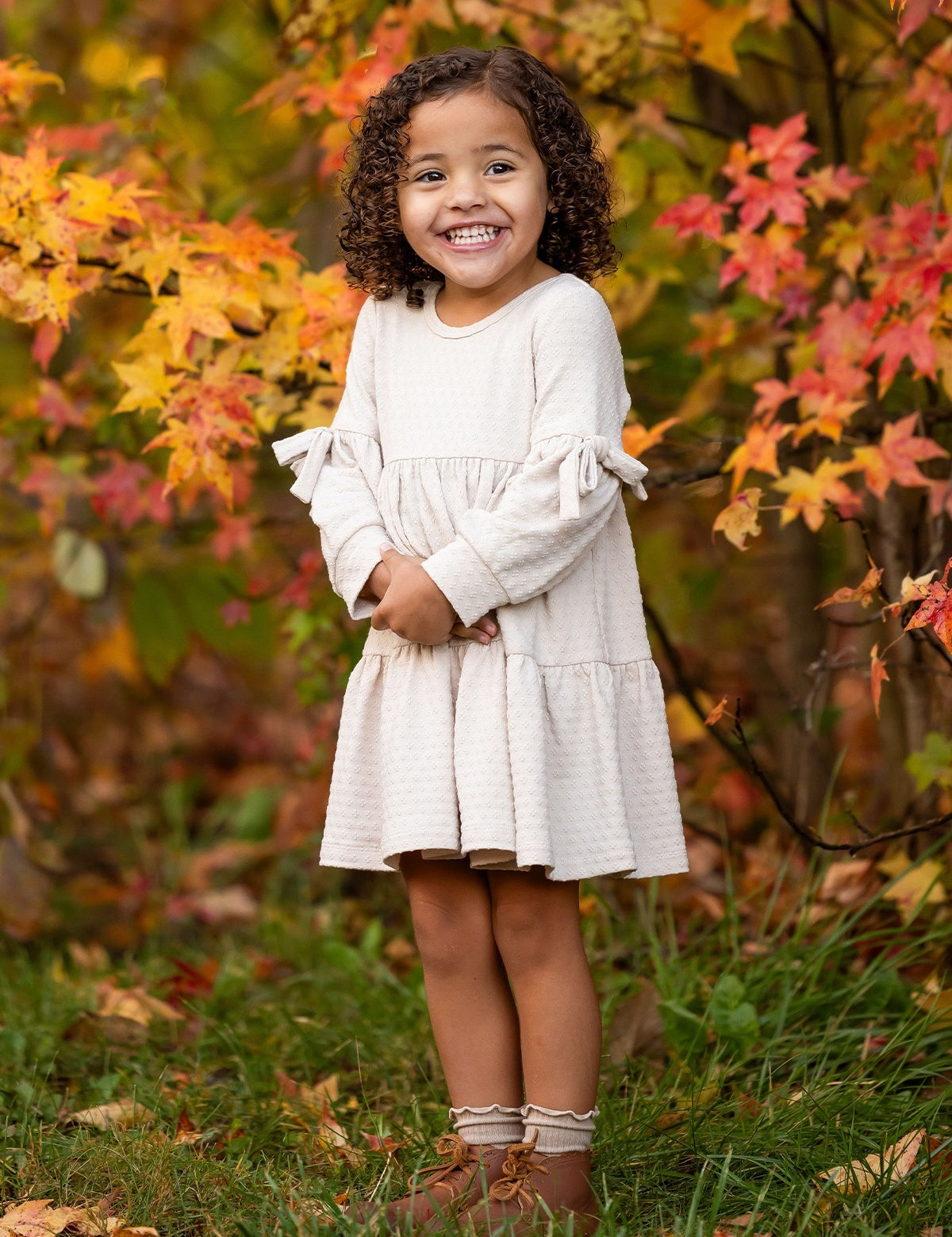 A young girl with curly hair smiles outdoors amid autumn leaves, wearing the Mabel and Honey Cinnamon Sugar Dress with sleeve bows and a tiered silhouette, paired with brown boots as she stands on green grass.