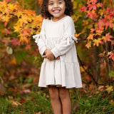 A young girl with curly hair smiles outdoors amid autumn leaves, wearing the Mabel and Honey Cinnamon Sugar Dress with sleeve bows and a tiered silhouette, paired with brown boots as she stands on green grass.