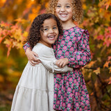 Two smiling girls with curly hair hug outdoors among vibrant autumn leaves. One wears a cream dress, the other a pink and green Mabel and Honey Cinnamon Sugar Dress featuring a playful tiered silhouette.
