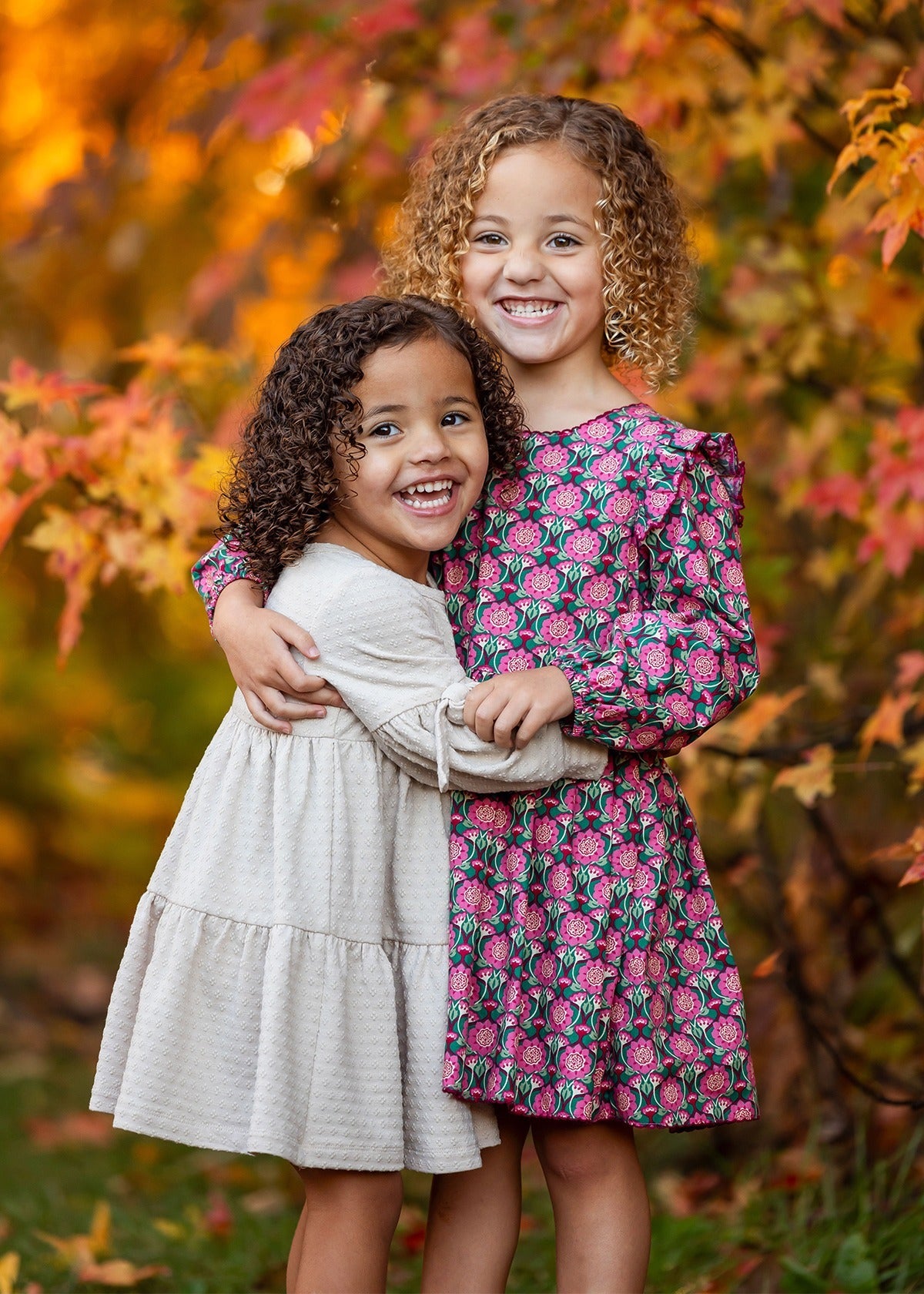 Two smiling girls with curly hair hug outdoors among vibrant autumn leaves. One wears a cream dress, the other a pink and green Mabel and Honey Cinnamon Sugar Dress featuring a playful tiered silhouette.