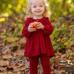 A smiling toddler with curly blonde hair stands outdoors on fallen leaves, wearing the Mabel and Honey Rosie Two Piece Set with ribbed leggings and a ruffled collar, holding a red apple against a blurred green and yellow foliage background.