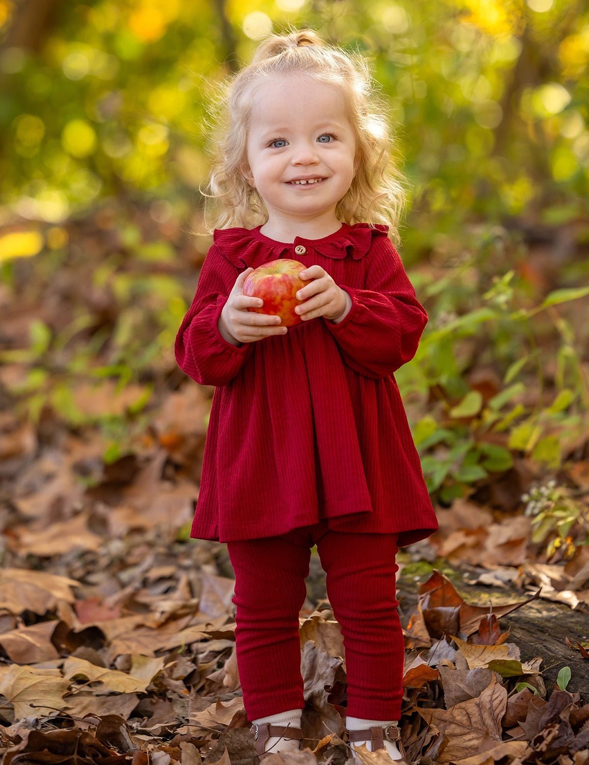 A smiling toddler with curly blonde hair stands outdoors on fallen leaves, wearing the Mabel and Honey Rosie Two Piece Set with ribbed leggings and a ruffled collar, holding a red apple against a blurred green and yellow foliage background.