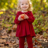 A smiling toddler with curly blonde hair stands outdoors on fallen leaves, wearing the Mabel and Honey Rosie Two Piece Set with ribbed leggings and a ruffled collar, holding a red apple against a blurred green and yellow foliage background.