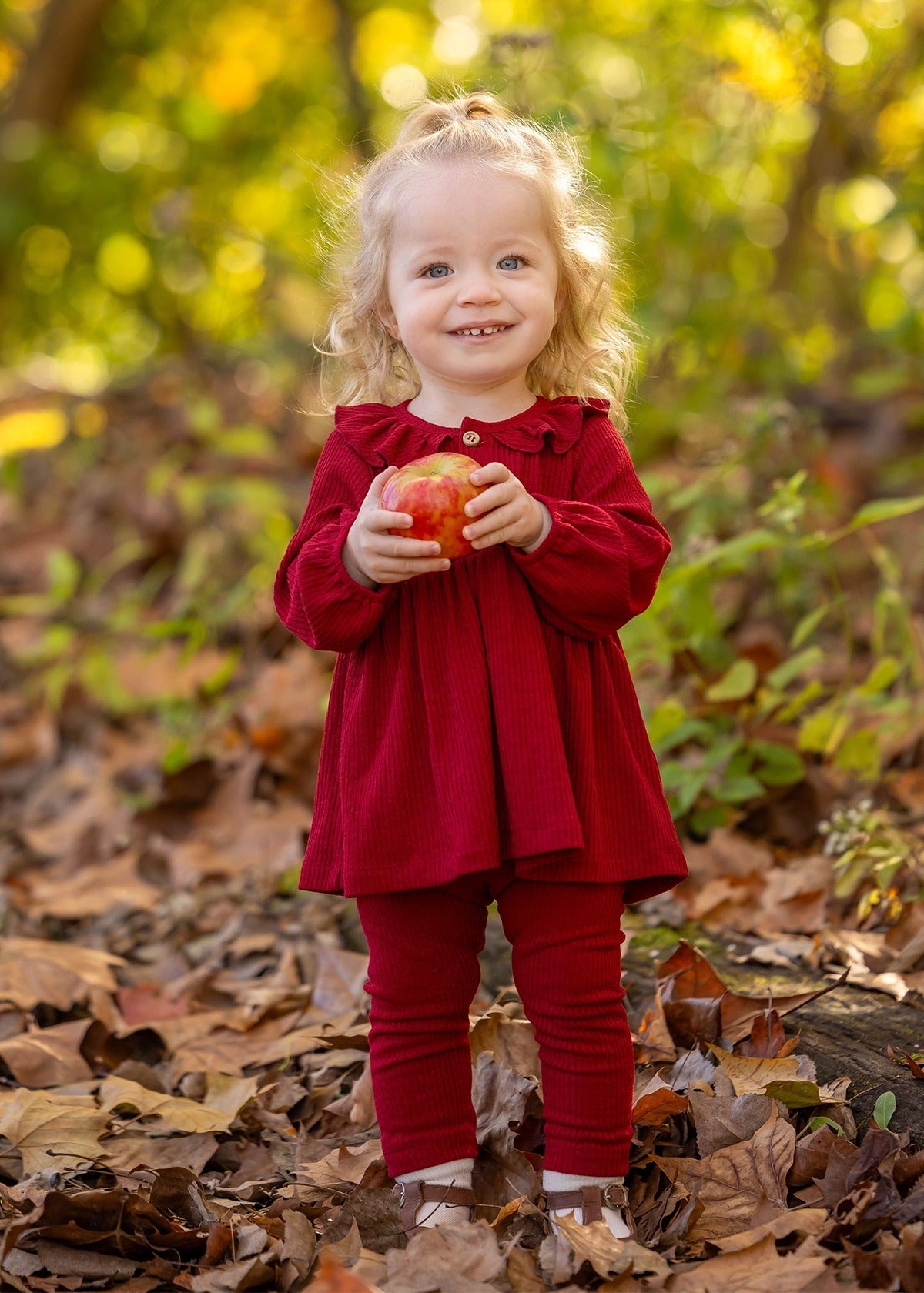 A smiling toddler with curly blonde hair stands outdoors on fallen leaves, wearing the Mabel and Honey Rosie Two Piece Set with ribbed leggings and a ruffled collar, holding a red apple against a blurred green and yellow foliage background.