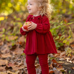 A smiling toddler in the Rosie Two Piece Set by Mabel and Honey stands among autumn leaves outdoors, holding a red apple with both hands. Green and yellow foliage is visible in the background.