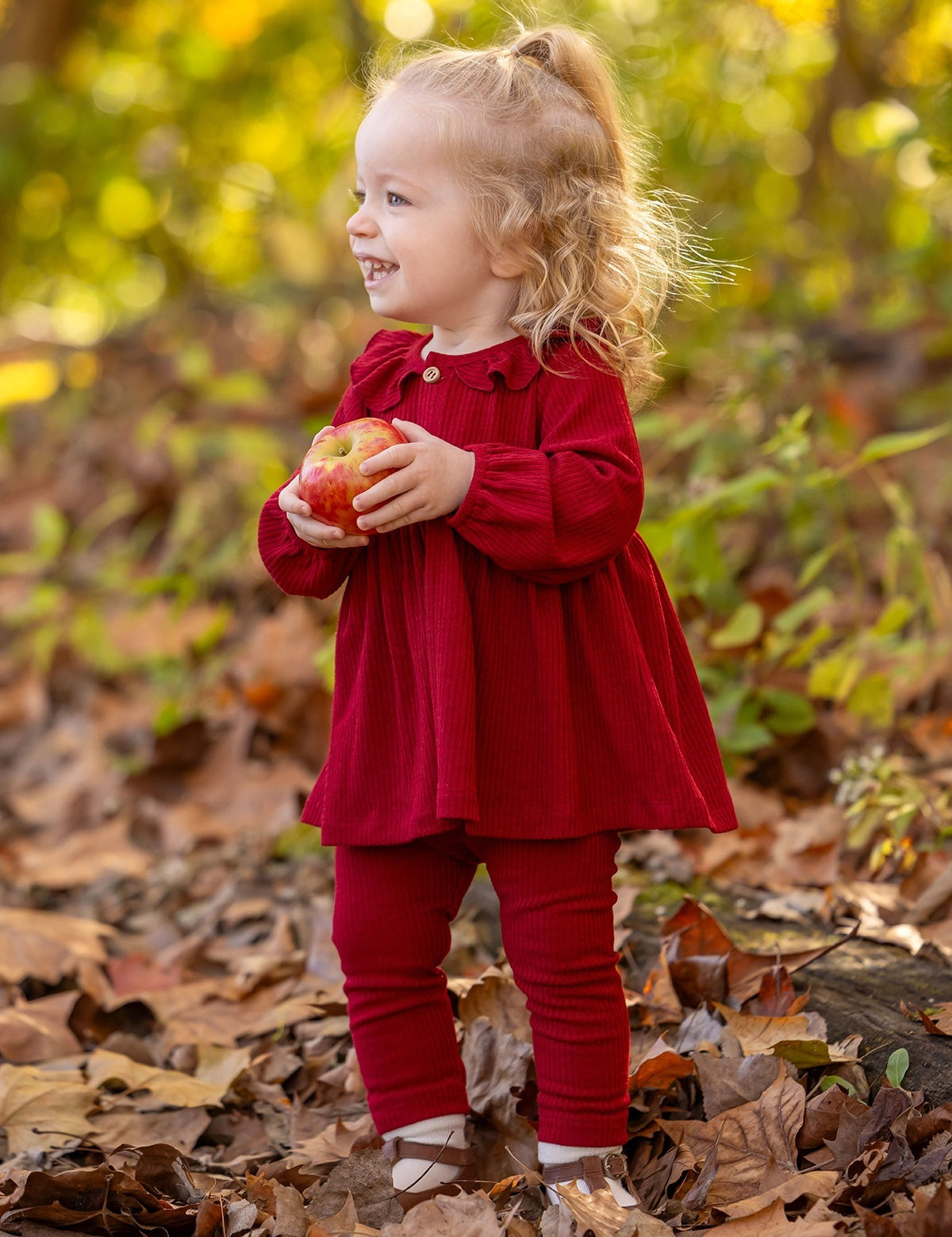 A smiling toddler in the Rosie Two Piece Set by Mabel and Honey stands among autumn leaves outdoors, holding a red apple with both hands. Green and yellow foliage is visible in the background.