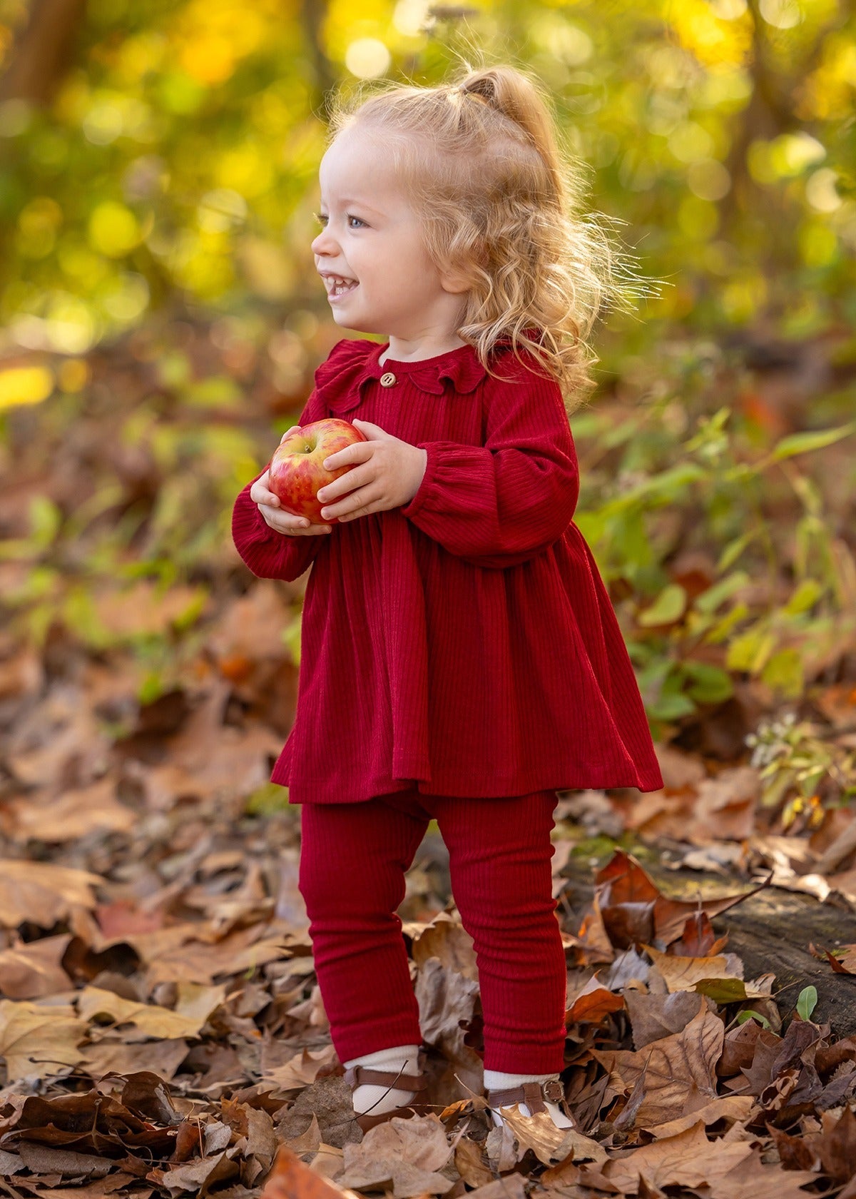 A smiling toddler in the Rosie Two Piece Set by Mabel and Honey stands among autumn leaves outdoors, holding a red apple with both hands. Green and yellow foliage is visible in the background.